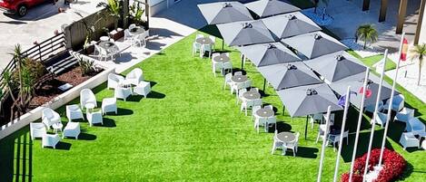 Un espace extérieur aménagé avec des tables et des parasols, entouré d’une pelouse bien entretenue et d’un bâtiment sur lequel on peut lire l’inscription « HARPZUL ».