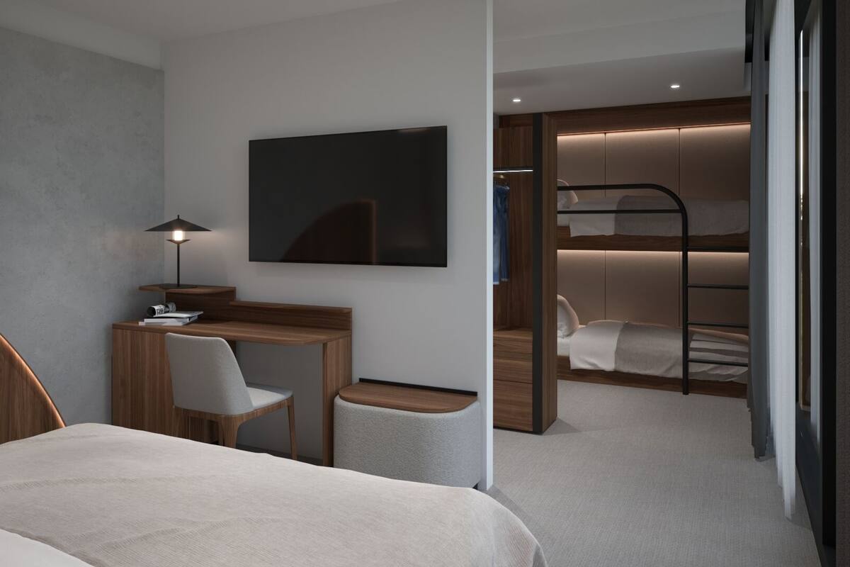 Quarto de hotel moderno com beliche, escrivaninha com cadeira, televisão de tela plana e uma pequena mesa de cabeceira.