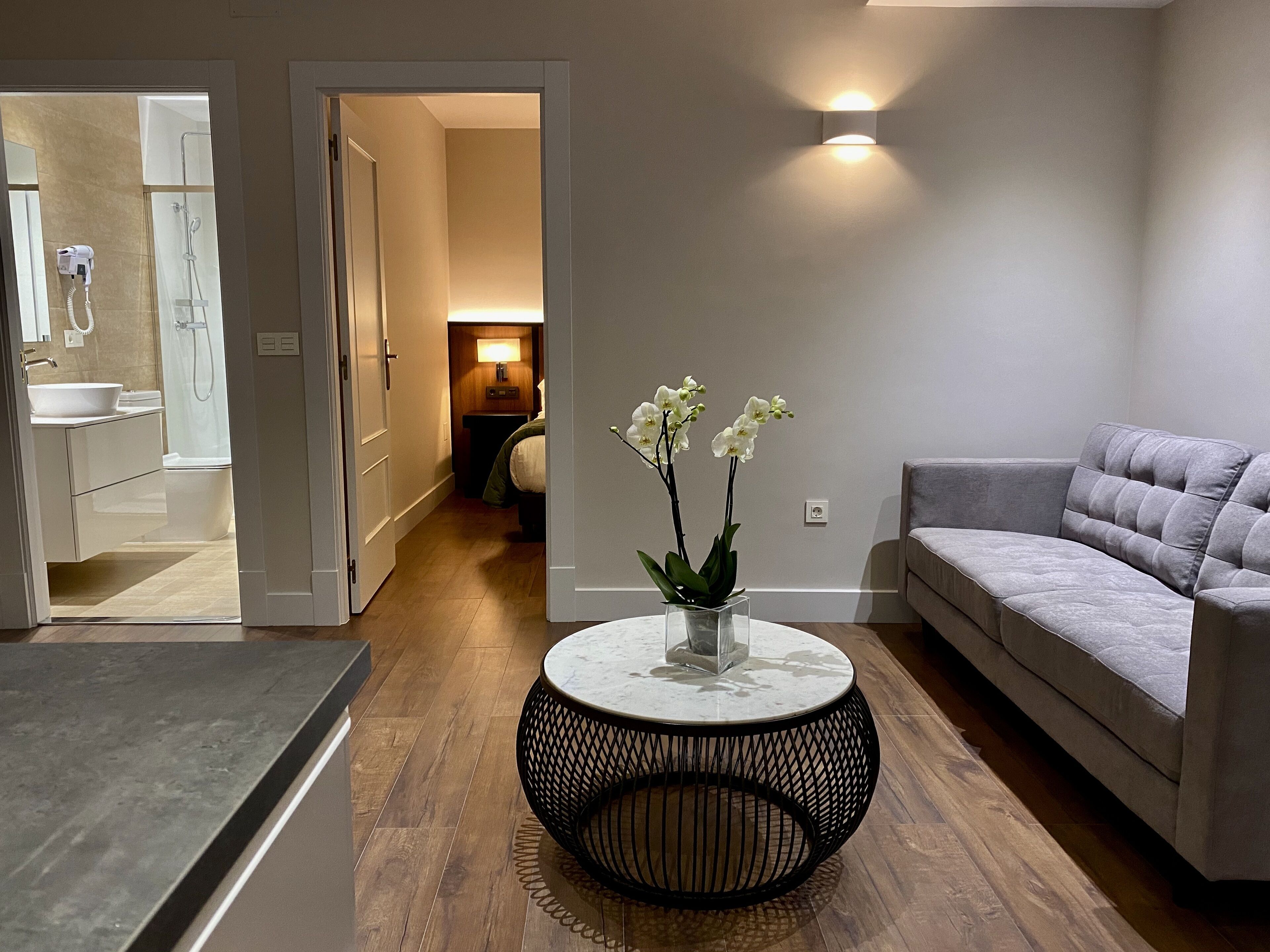 Een moderne woonkamer met een grijze bank, een ronde salontafel met een vaas orchideeën, en een gang die naar een badkamer leidt.