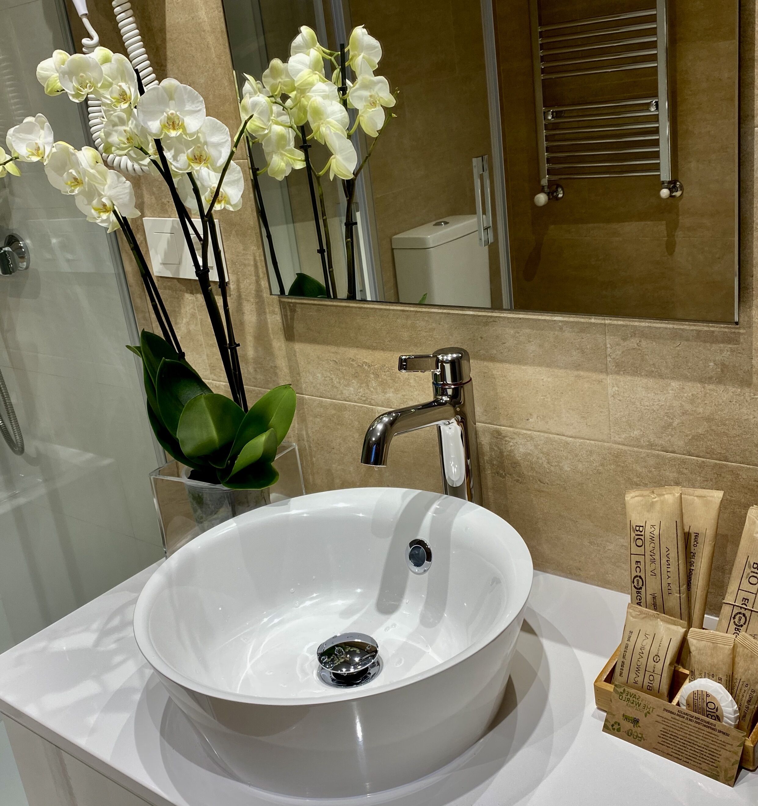 Une salle de bain avec un lavabo blanc, un miroir et des toilettes. Un vase contenant des orchidées blanches est posé sur le comptoir.