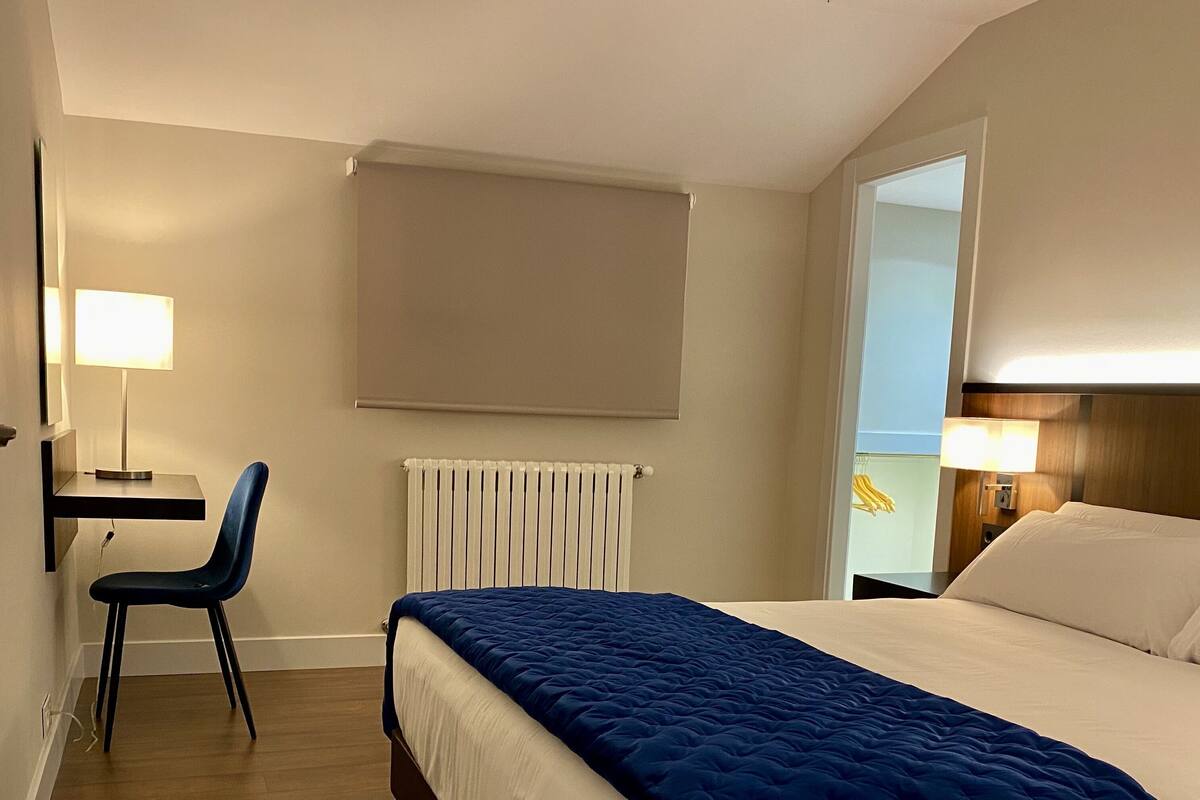 Quarto de hotel com cama, cadeira, escrivaninha, radiador e janela.