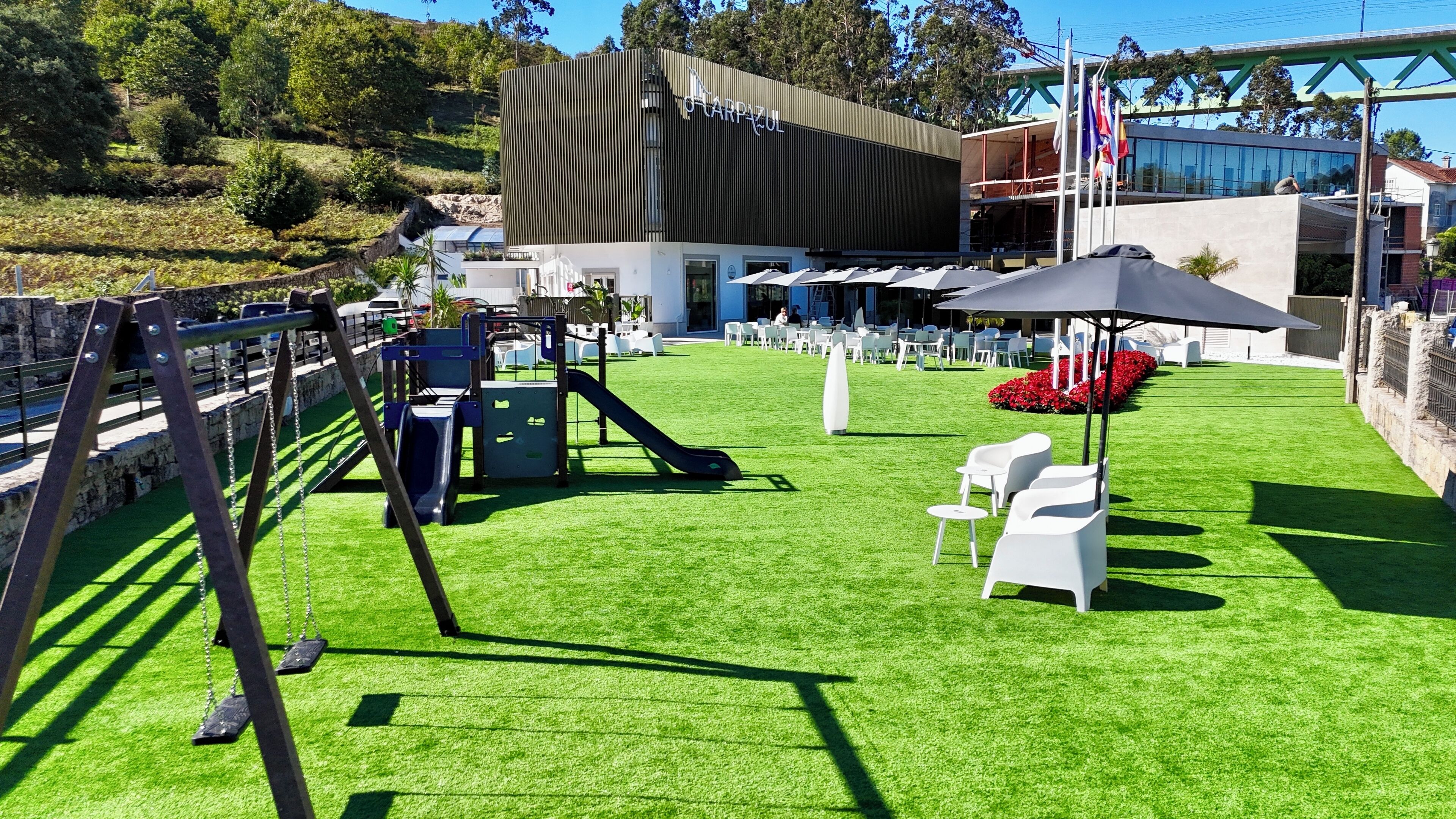 A playground with slides and swings, a seating area with white chairs and a table, and a building with a modern design.