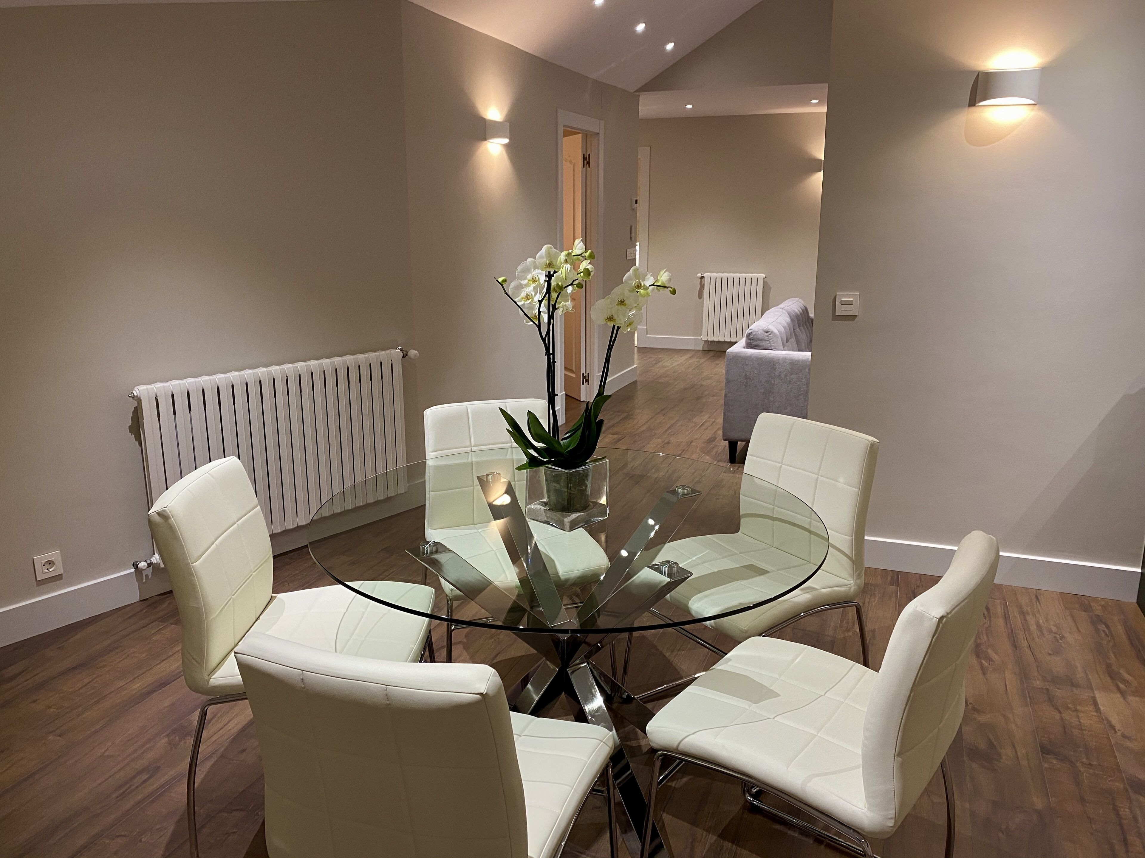 A modern dining area with a glass table surrounded by white chairs, a vase with flowers, and a white radiator on the wall.