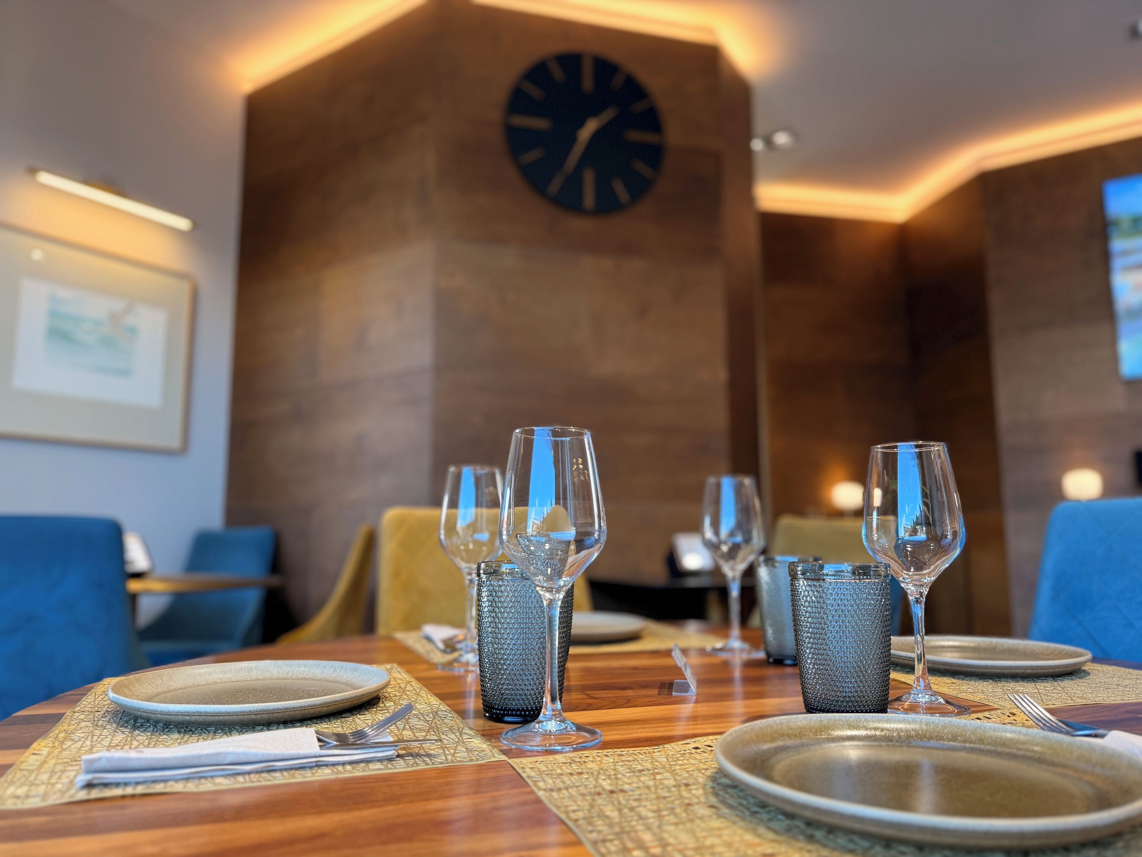 A dining table set with plates, cutlery, and glasses, with a clock on the wall and a framed picture in the background.