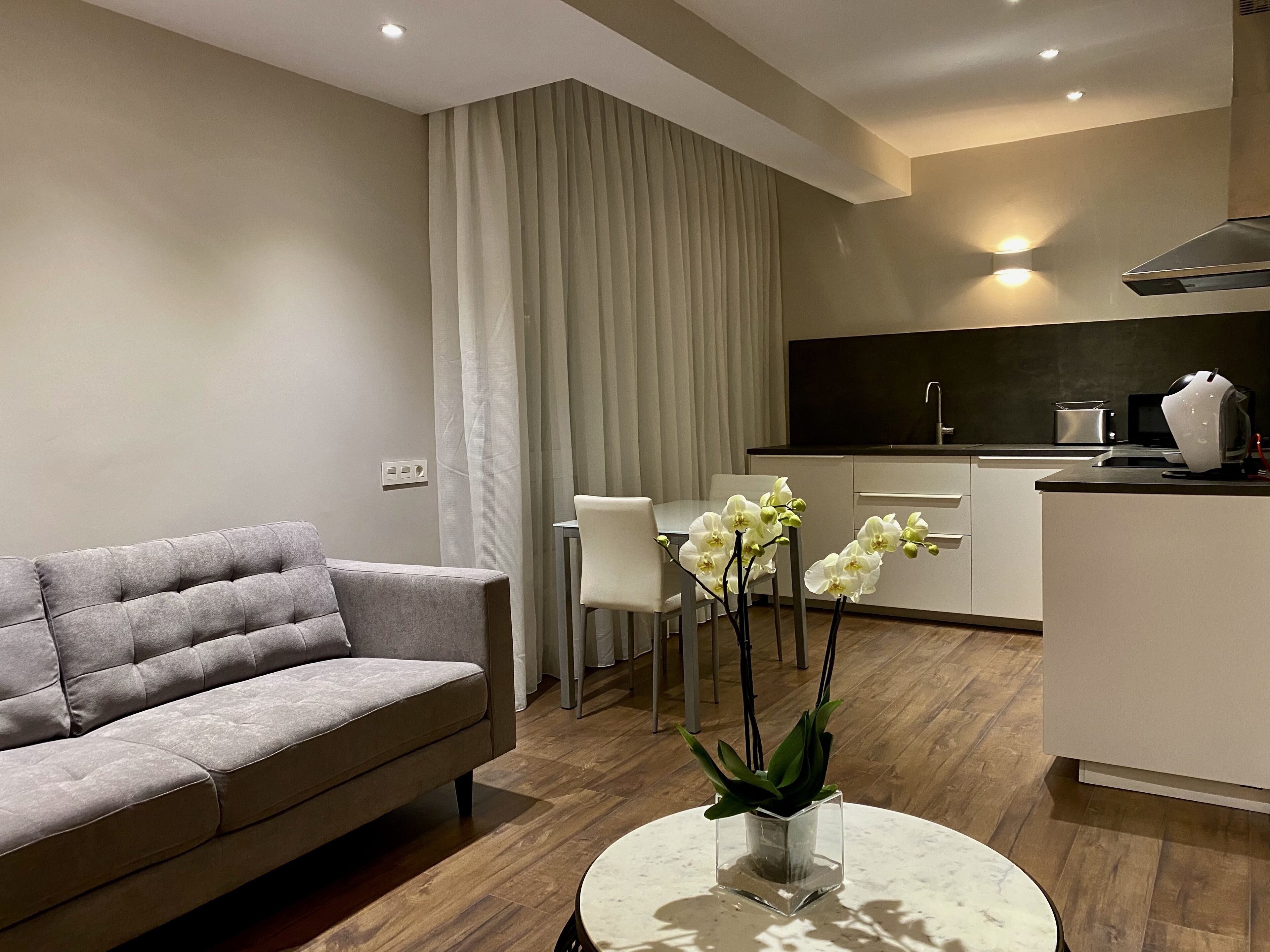 A modern living room with a grey sofa, a dining table with chairs, a small white table with orchids, and a kitchen area with a stove and microwave.