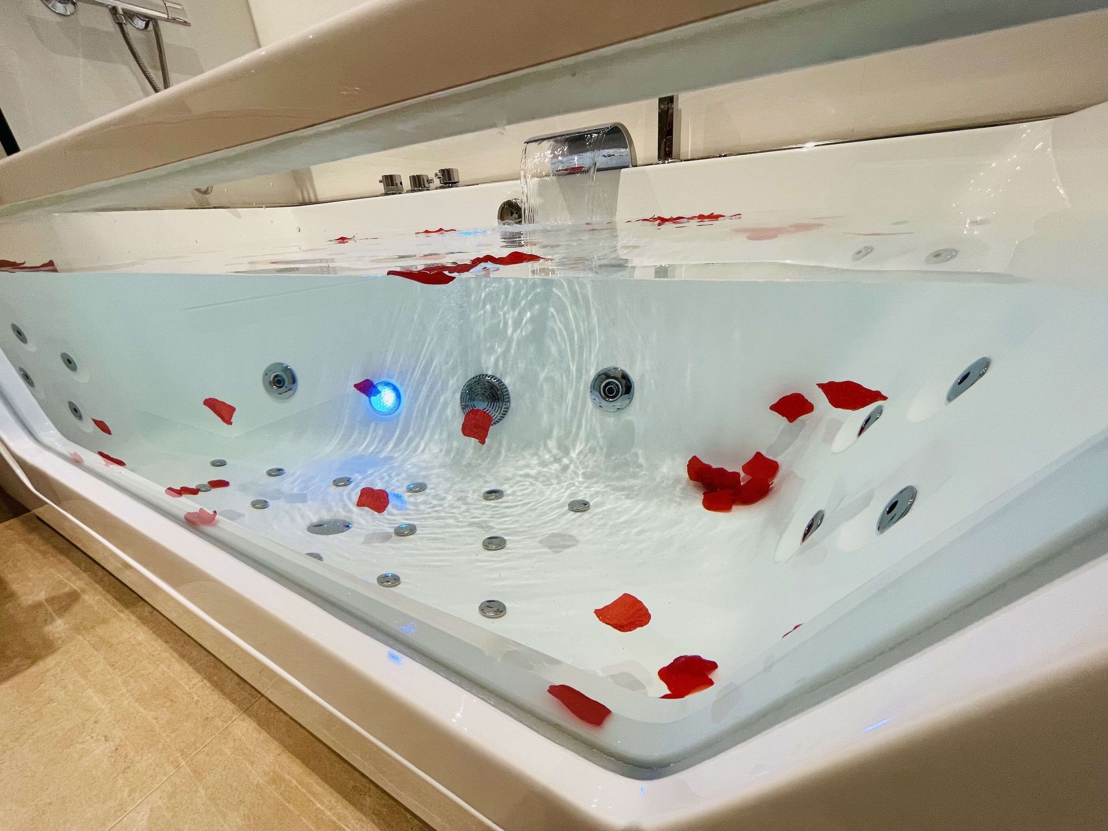 Une salle de bain moderne avec une grande baignoire remplie d’eau et de pétales de rose, dotée de plusieurs jets et d’un éclairage intégré.
