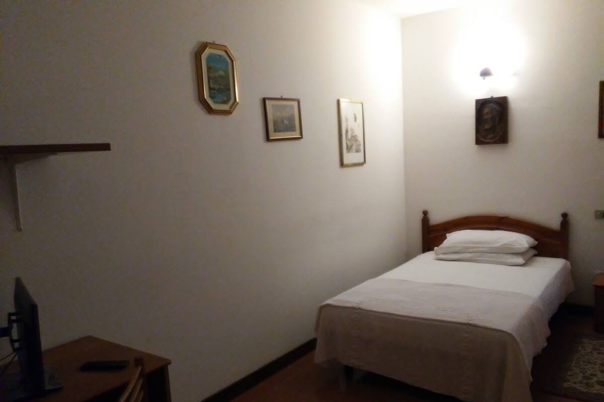 A bedroom with a single bed, a desk with a computer, and a chair. There are framed pictures on the wall.