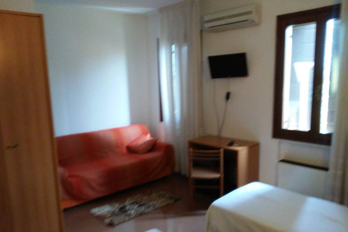A room with a red sofa, a small desk, a bed, and a TV mounted on the wall.