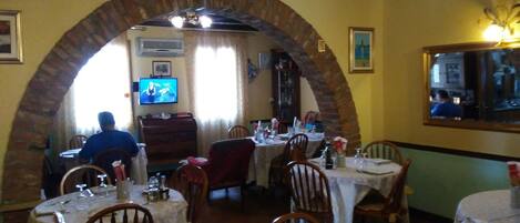Um restaurante com entrada em tijolos com arco, mesas postas para refeições e uma televisão ao fundo.