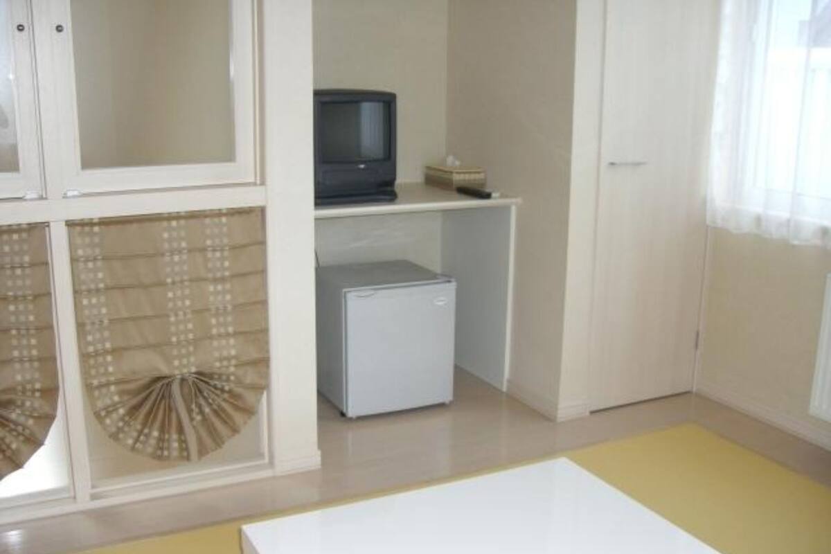 A room with a white table, a small white refrigerator, a television on a shelf, and a window with sheer curtains.
