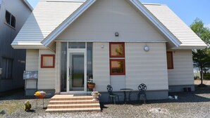A single-story building with a gabled roof, a front door, and a small patio area with two chairs and a table. - Meguminoyu (Furano)