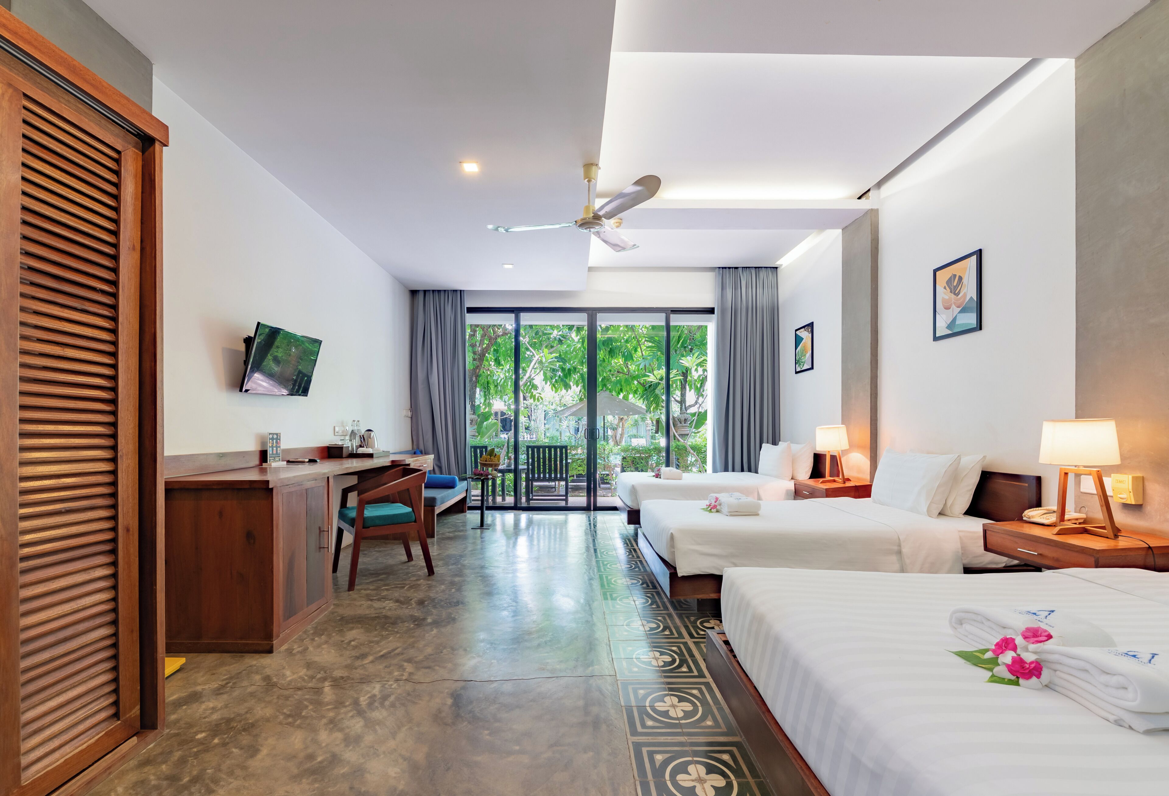 A hotel room with a desk, two beds, a ceiling fan, and a view of greenery outside.