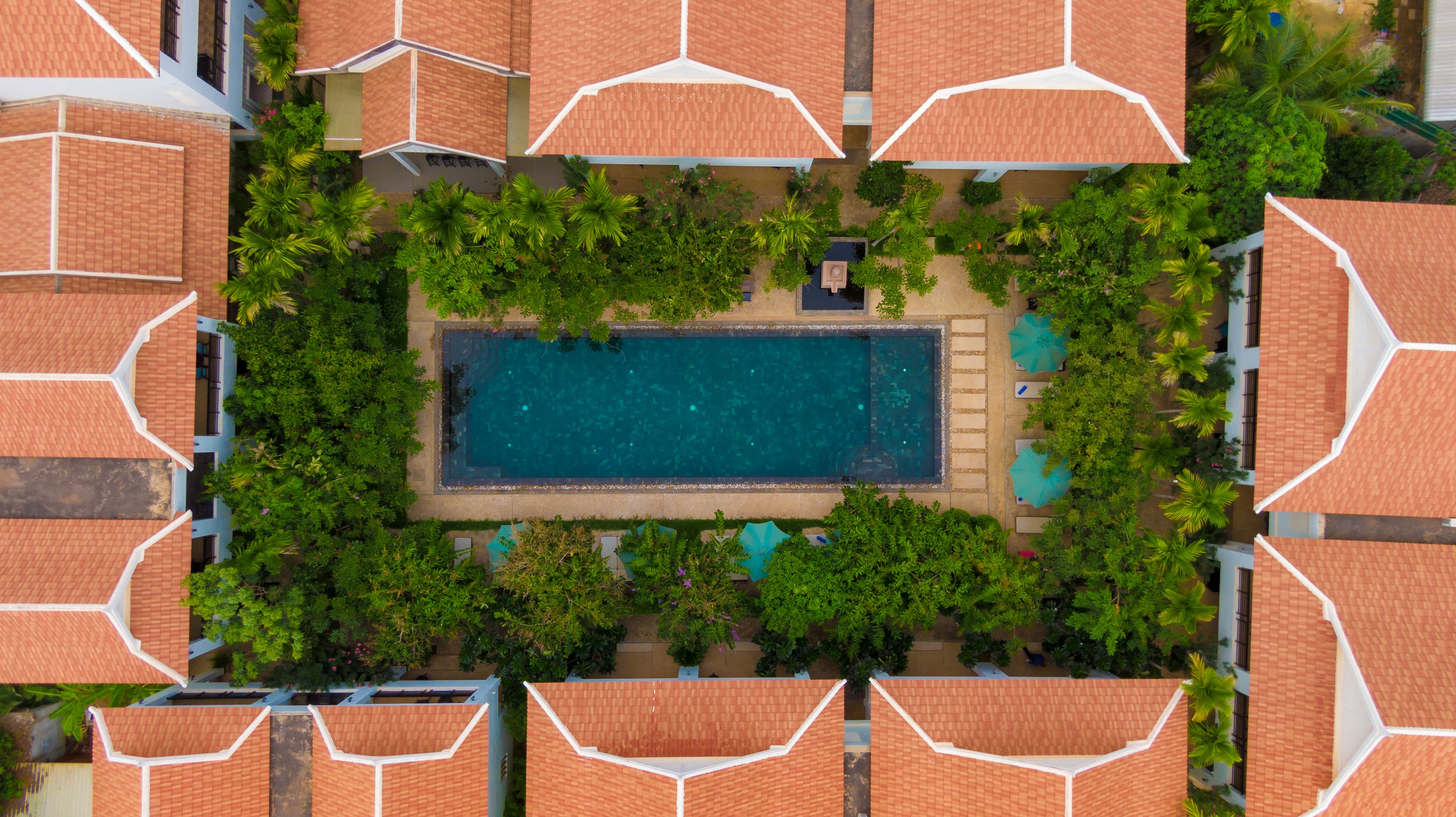Vue aérienne d’un complexe hôtelier doté d’une piscine centrale, entourée d’une végétation luxuriante et de plusieurs structures dotées de toits.