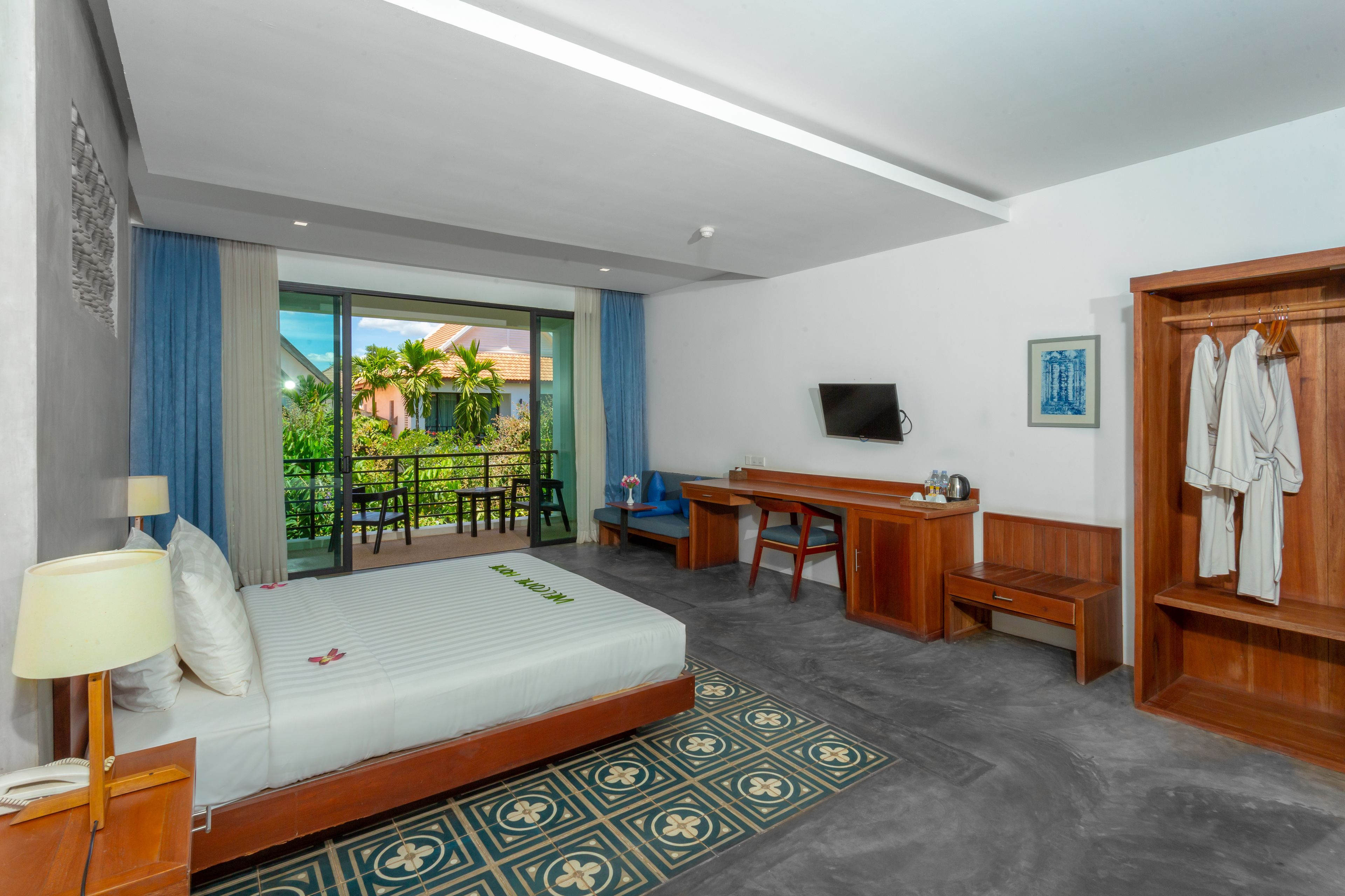 Habitación de hotel con cama, escritorio, silla y televisor. Tiene un balcón con vistas a zonas verdes y edificios.