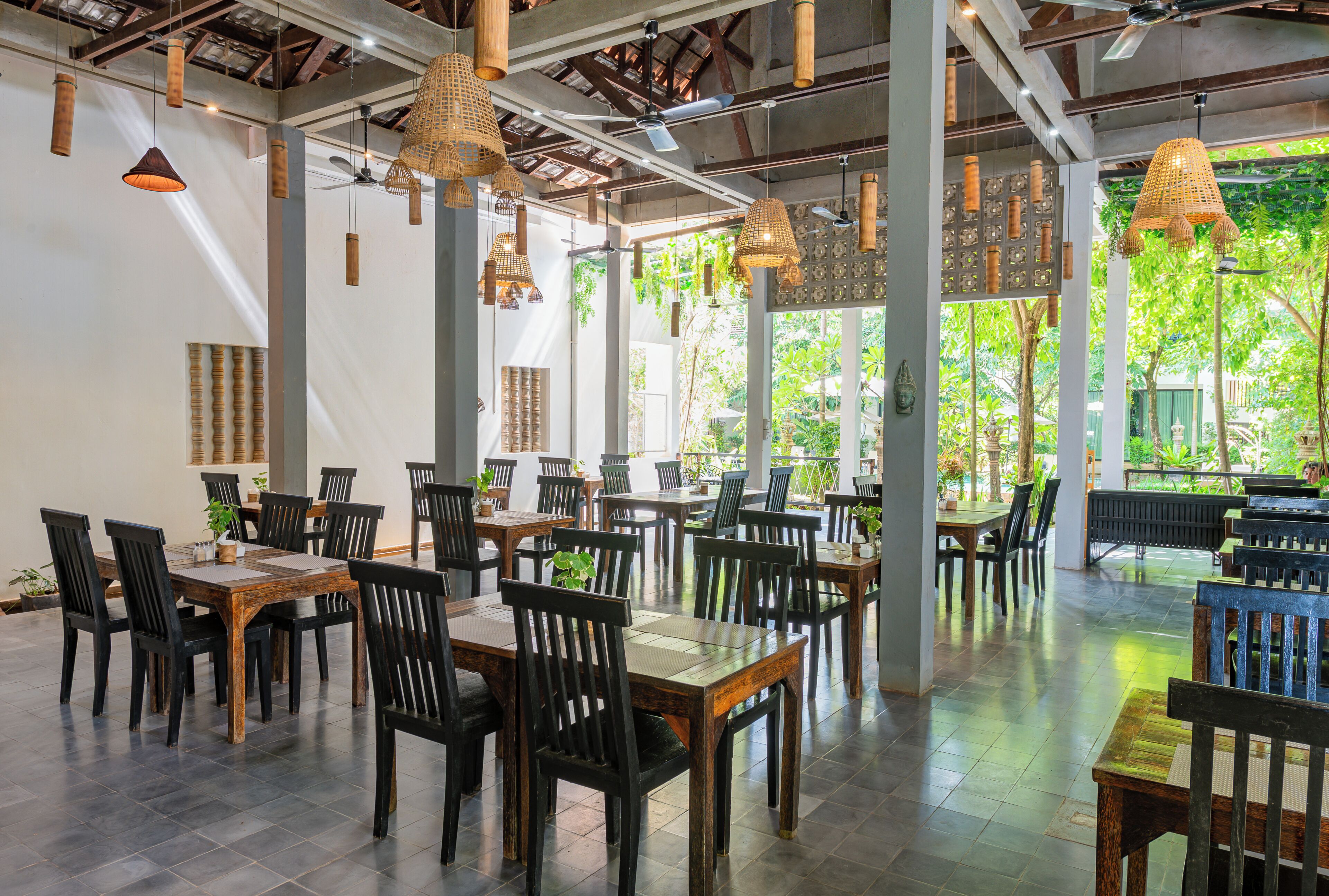 A spacious indoor dining area with multiple wooden tables and chairs, large windows allowing natural light, and hanging wicker lamps.