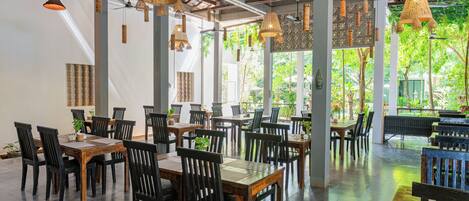 A spacious indoor dining area with multiple wooden tables and chairs, large windows allowing natural light, and hanging wicker lamps.