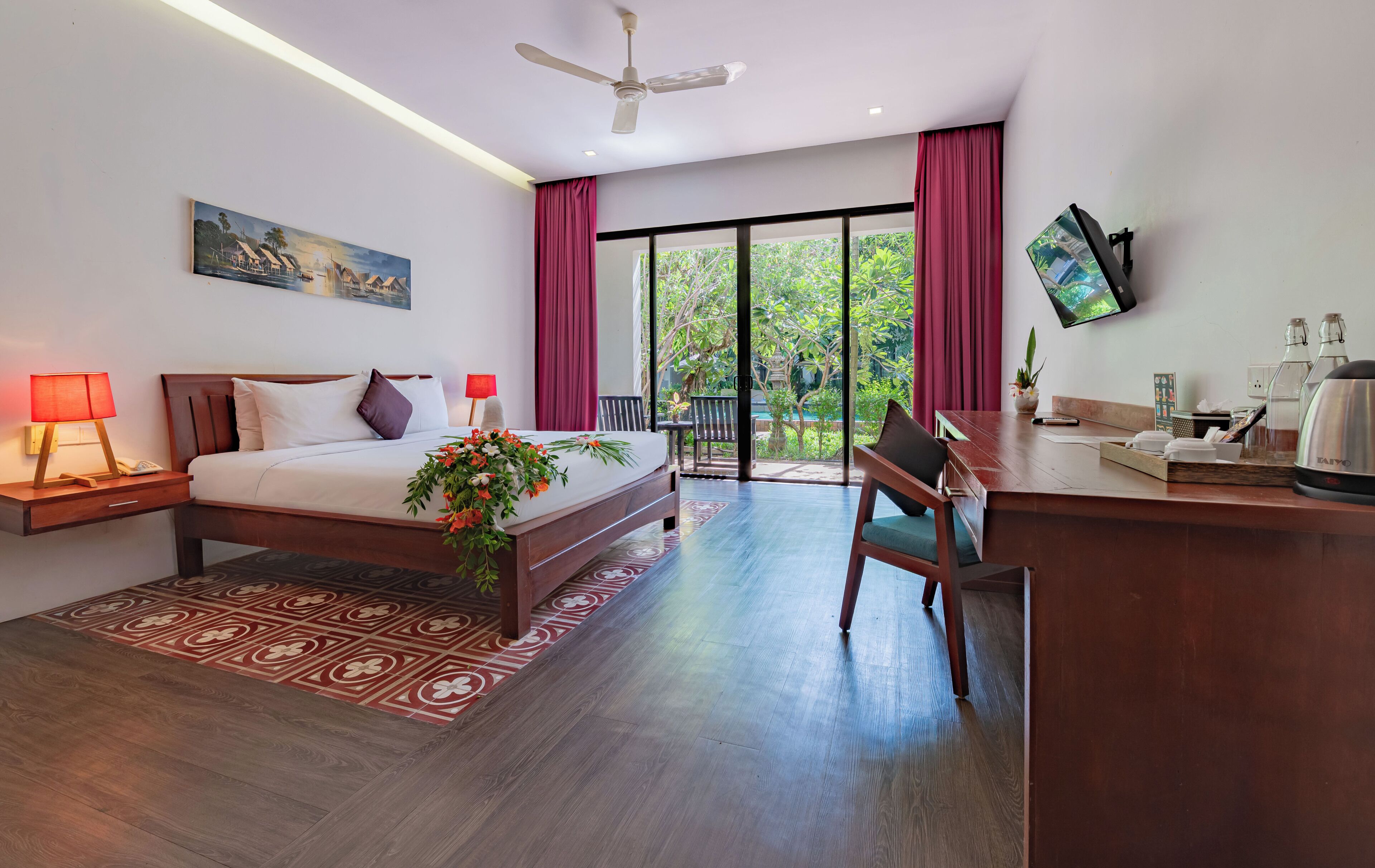 A hotel room with a bed, bedside lamps, a desk, a television, and a view of greenery through the window.
