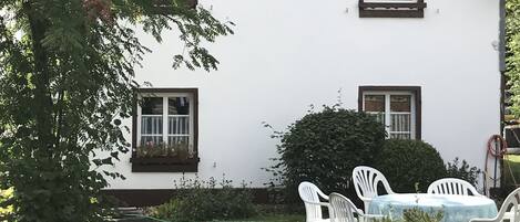 Una casa blanca con tejado oscuro, varias ventanas y un pequeño jardín con sillas y una mesa blancos.