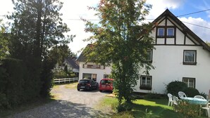 A two-story building with a white facade and dark trim. There are two cars parked in front, and a gravel path leading to the entrance. - Pension Falter (Nideggen)