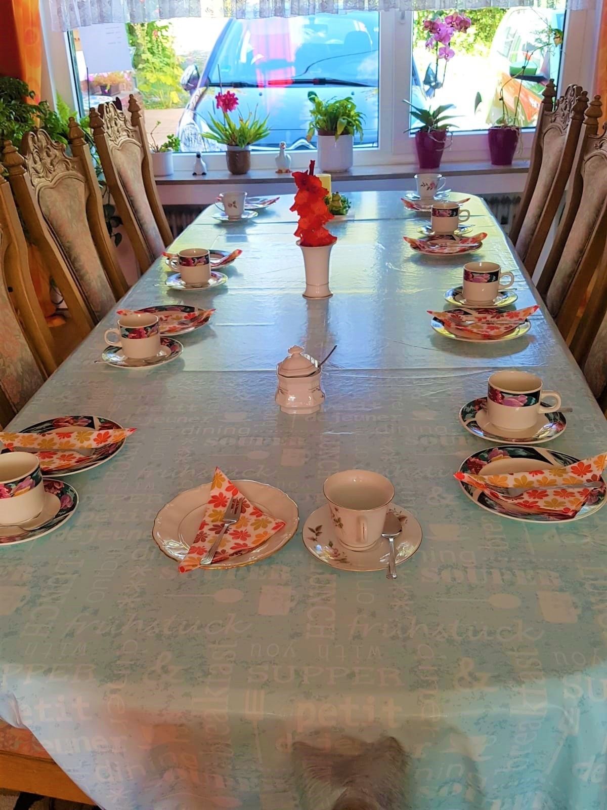 Uma mesa de jantar posta para uma refeição, com toalhas de mesa florais, cadeiras e um vaso com arranjo de flor vermelha.