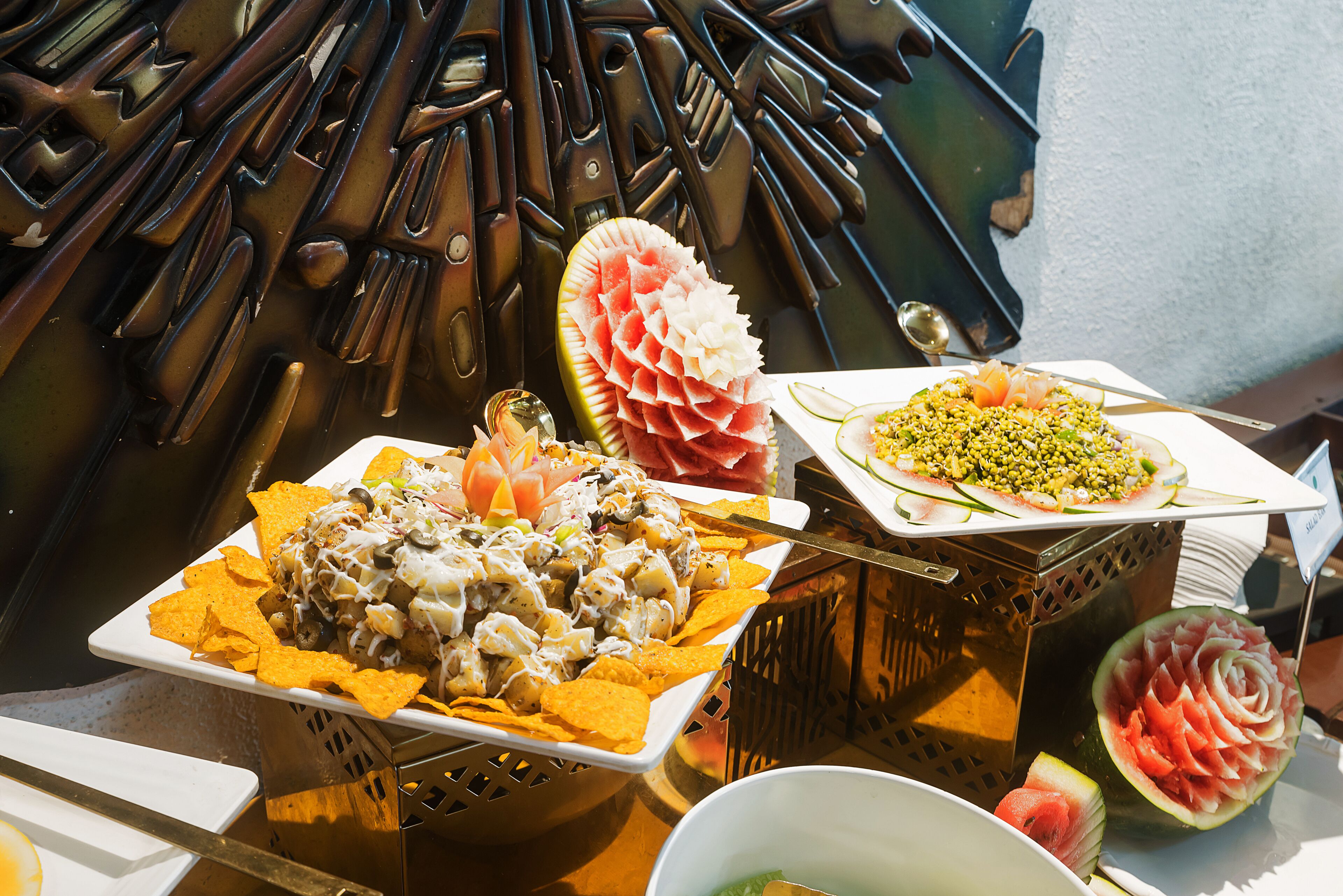 A buffet with nachos, a watermelon carved into a flower, and a plate of salad.