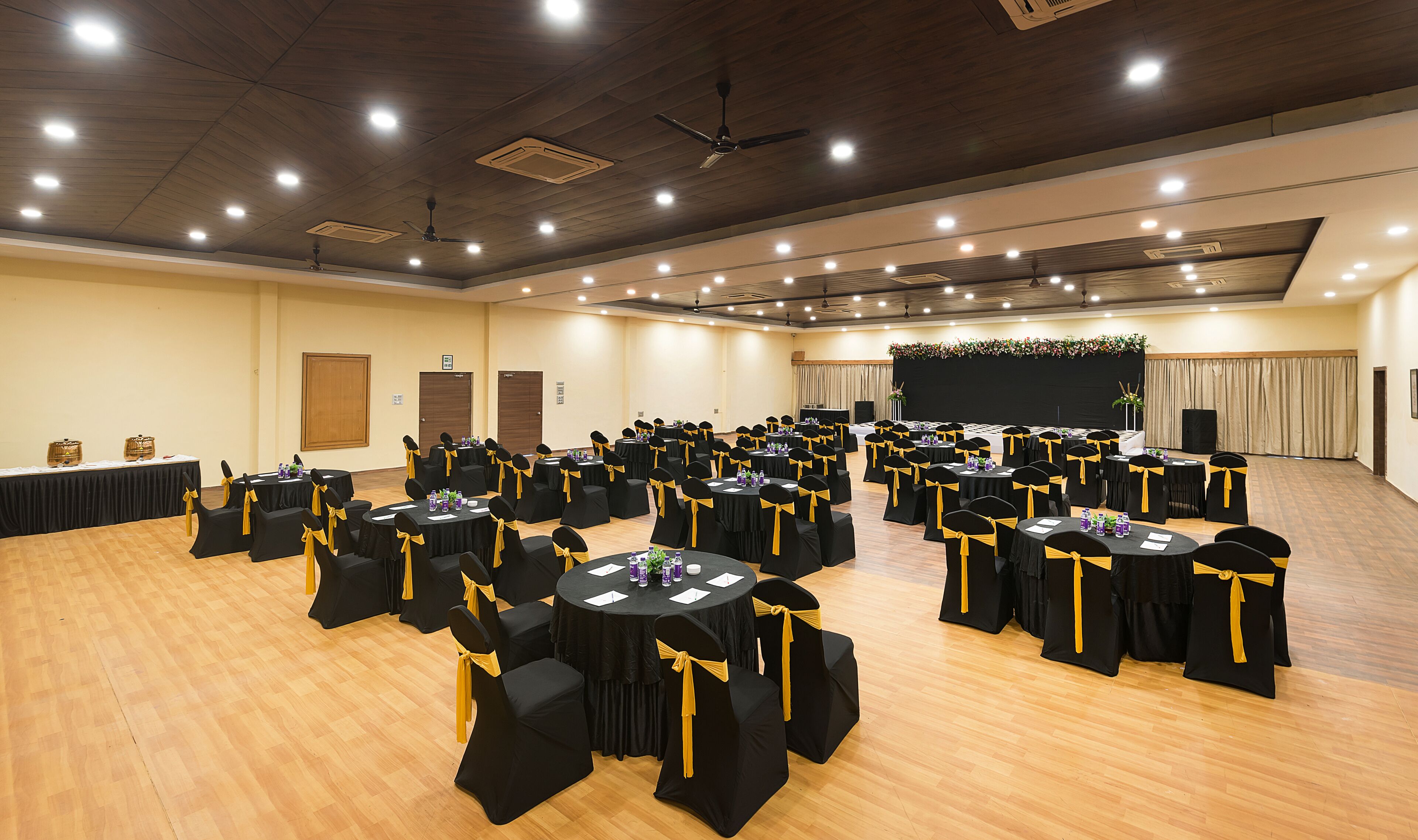 A conference room with multiple round tables, black chairs with yellow sashes, and a stage with a floral backdrop.
