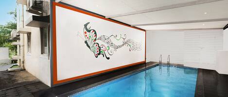 An indoor swimming pool with a decorative wall mural, a clear pool edge, and a tiled floor.