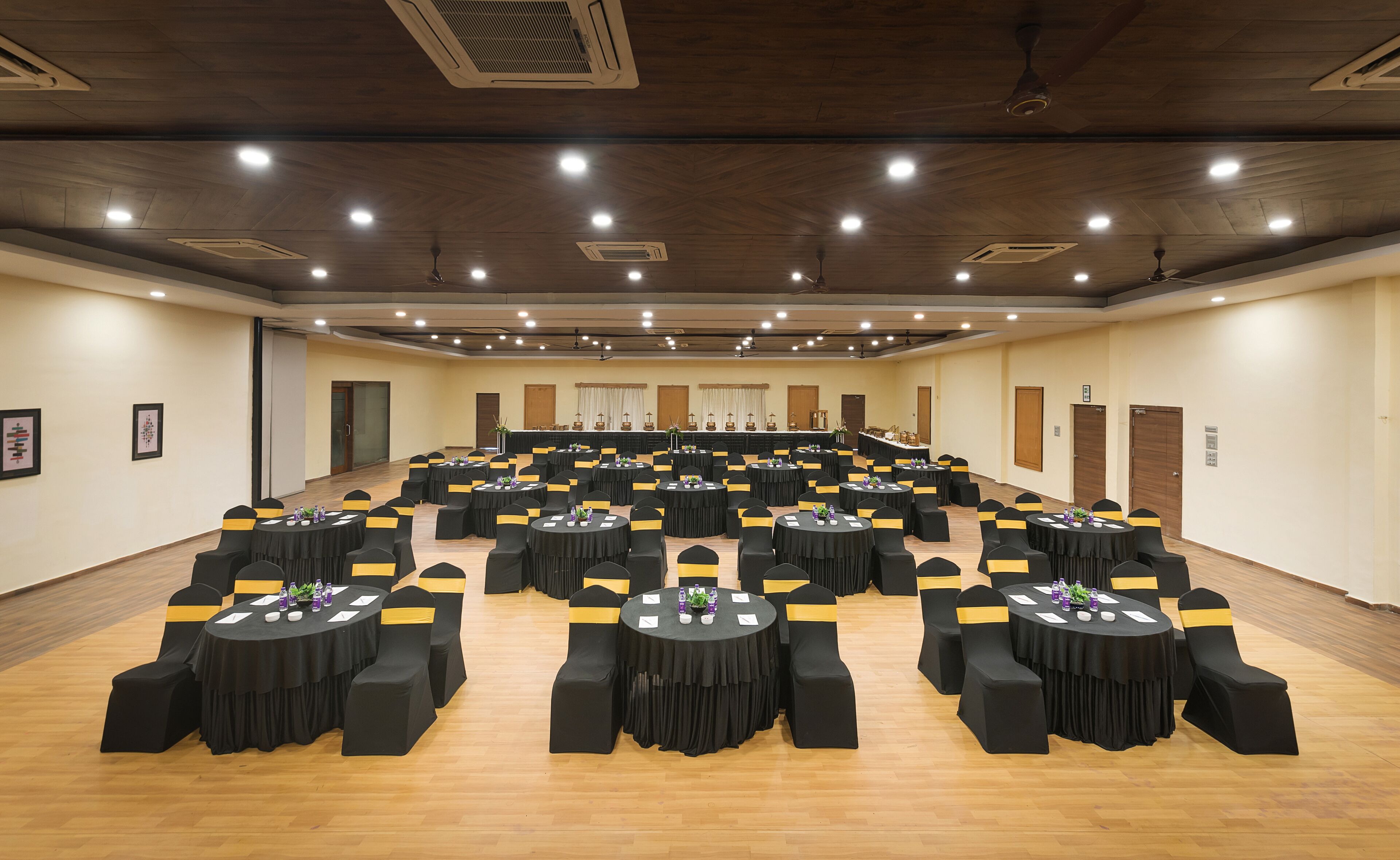 A large conference room with multiple round tables, black chairs with yellow sashes, and wooden floors.