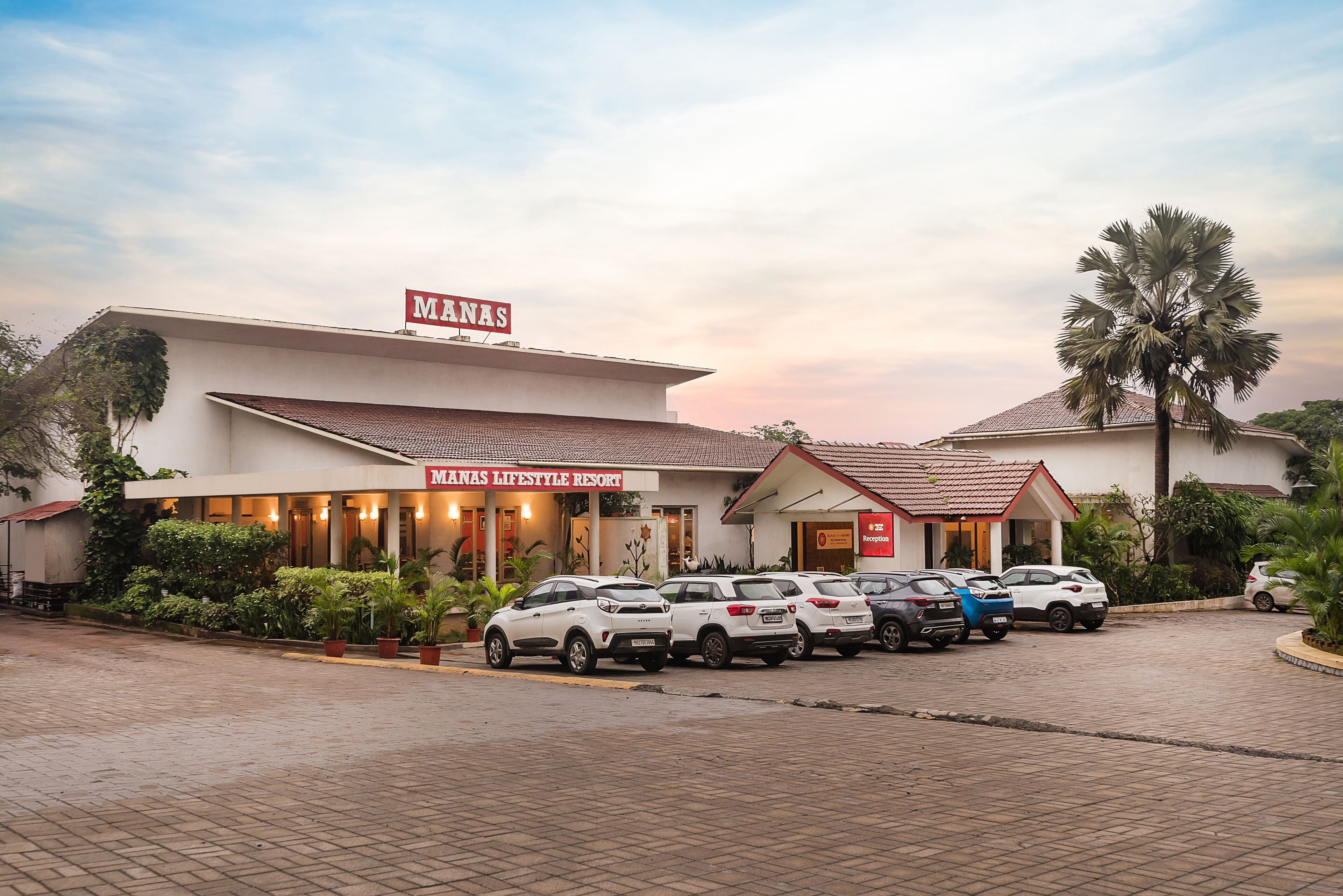 A building with a sign reading "MANAS LIFESTYLE RESORT" and a parking area with several cars.