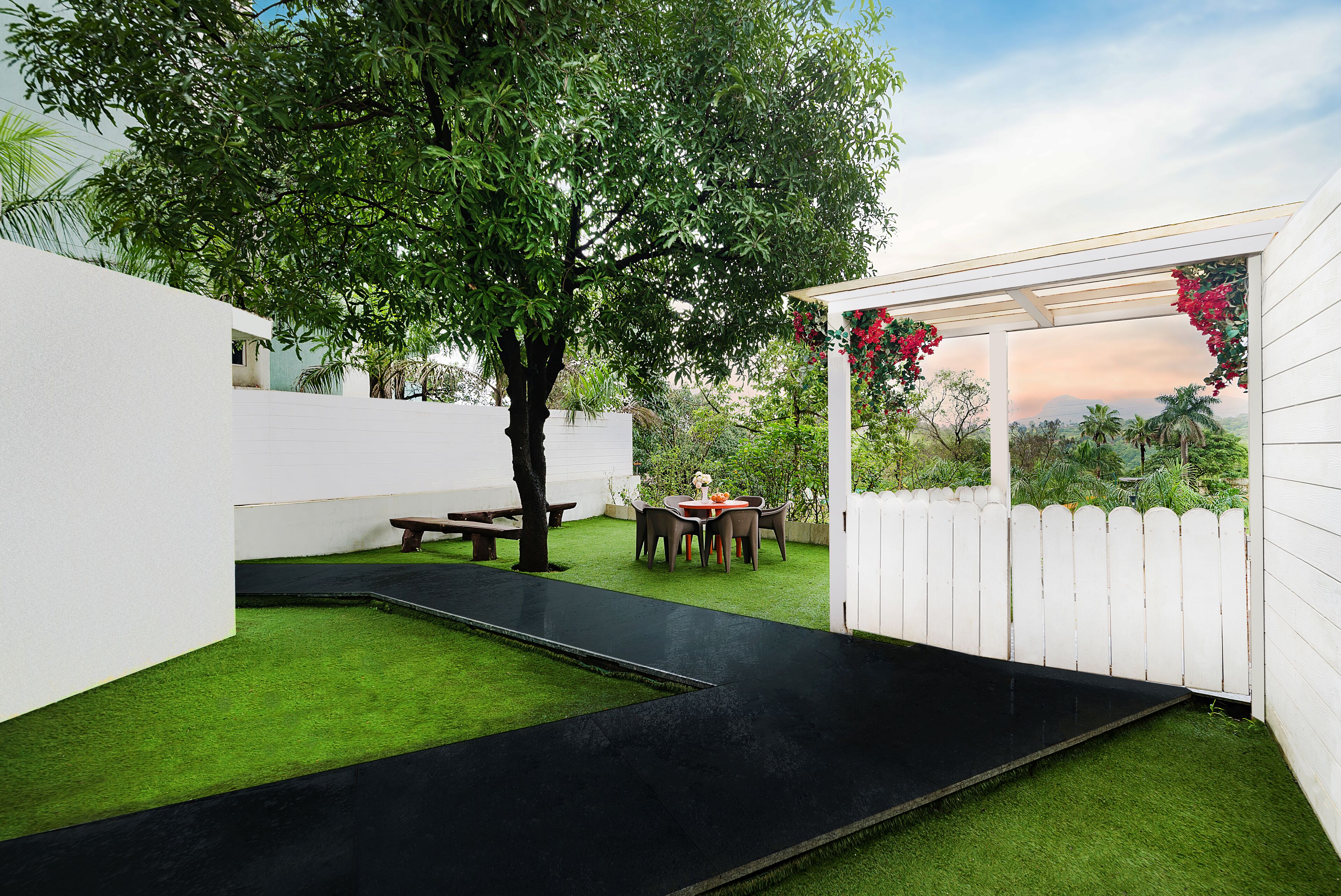 Patio esterno moderno con prato artificiale, vialetto nero, recinzione bianca e una pergola con fiori rossi.