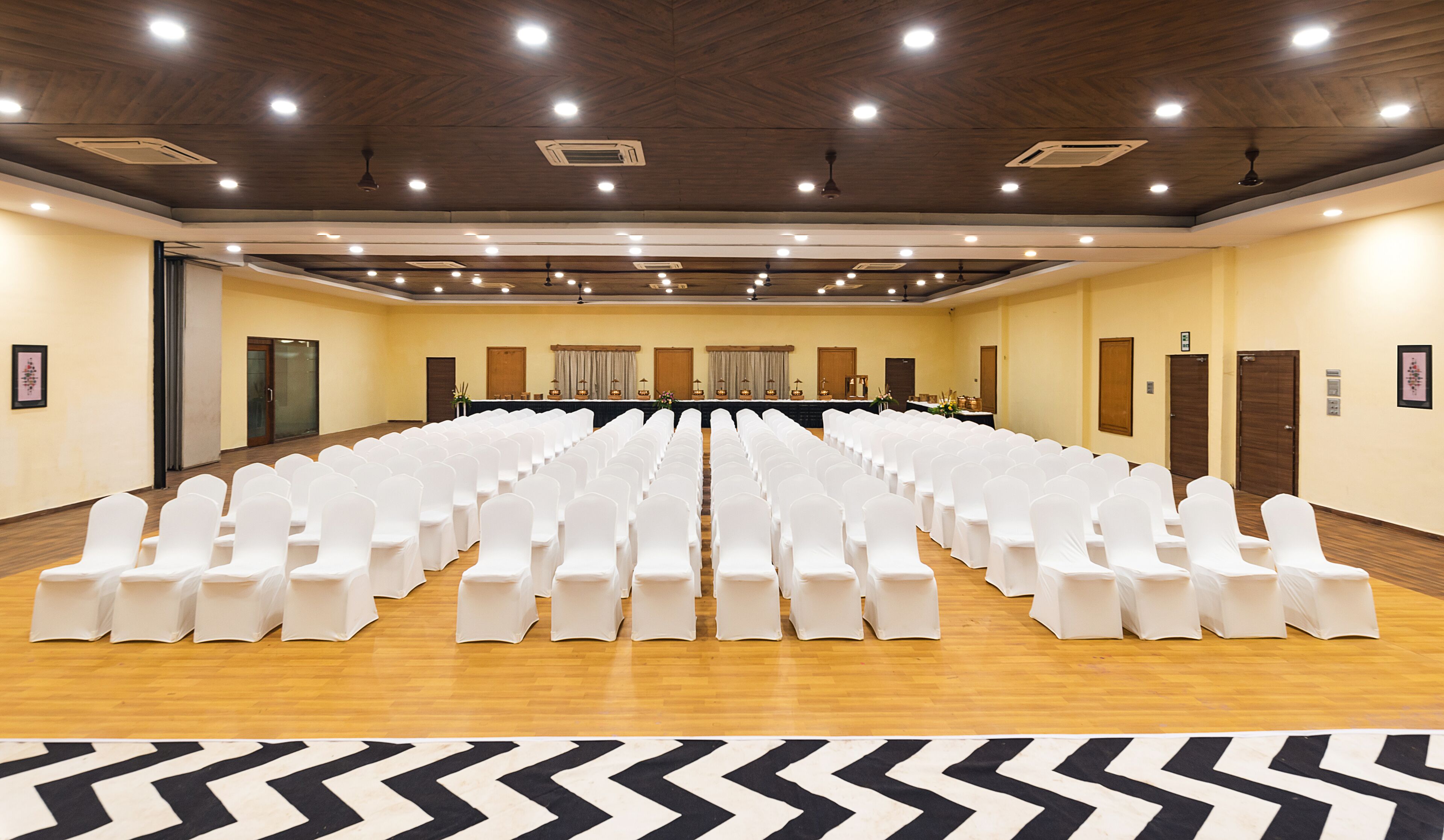 Un salón de conferencias con filas de sillas blancas, una mesa larga, suelo de madera y una alfombra estampada en patrón de rombos.