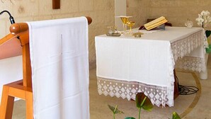 Interior de una capilla con una cruz en la pared, un altar blanco y una silla de madera cubierta con tela blanca.