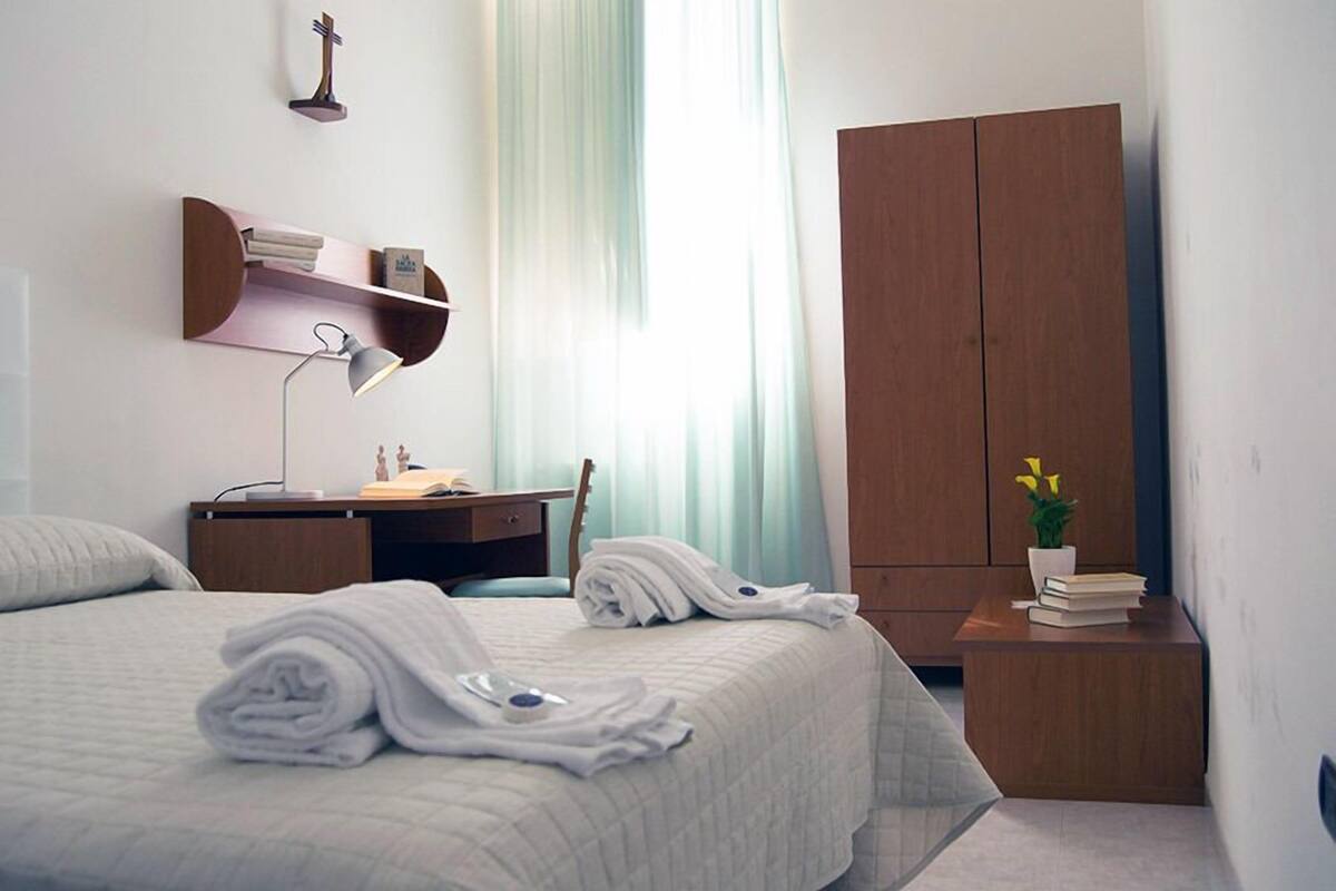 A neatly made bed with folded towels, a desk with a lamp, a wardrobe, and a small table with books and a plant.