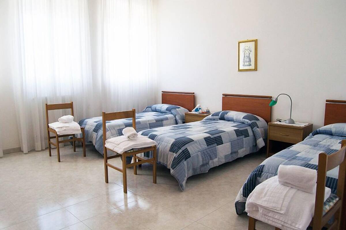 A hotel room with three single beds, each with a blue and white striped duvet, wooden headboards, and a bedside table with a lamp. There is a chair with a towel on it and a framed picture on the wall.