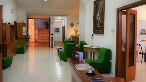 Un hall de hotel con mostrador de recepción, sillones verdes, una escalera y un cuadro en la pared.
