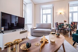 A modern living room with a flat-screen TV, grey sofa, floral chairs, and a wooden coffee table with decorative items.