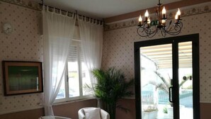A hotel room with a tiled floor, a white armchair, a small table, a potted plant, a chandelier, and a sliding glass door leading to a balcony. - Hotel Mon Reve (Rimini)
