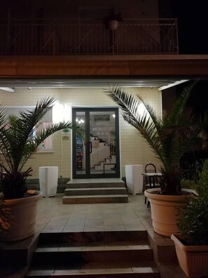 Entrada de un hotel con puerta de cristal, escalones que llevan a ella y plantas en macetas a ambos lados.