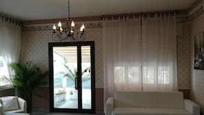 A living room with a white sofa, a small table, a potted plant, and a chandelier. There is a glass door leading to an outdoor area with a view of a building and palm trees. - Hotel Mon Reve (Rimini)