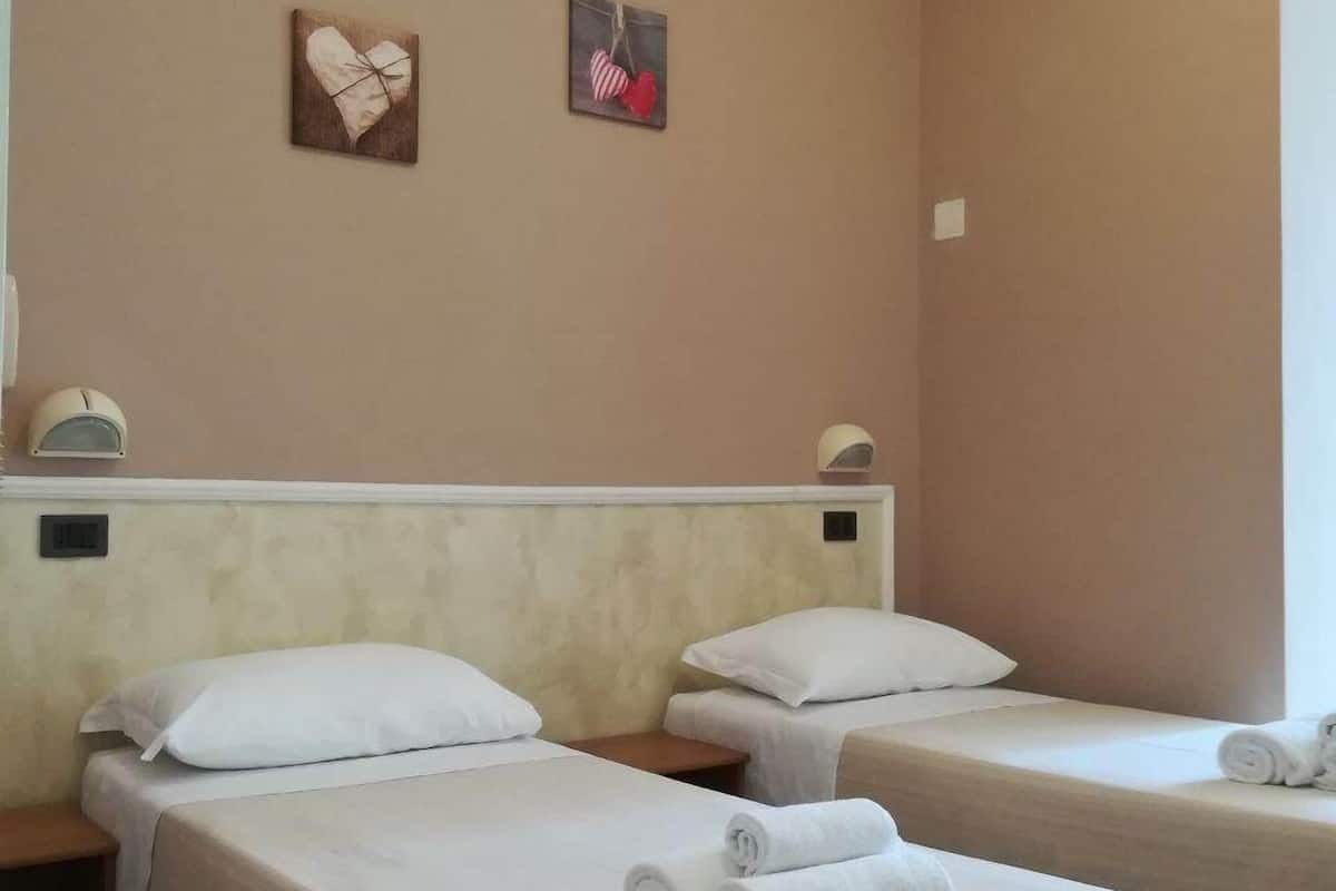 A hotel room with two beds, each with white linens and rolled towels. There are two wall-mounted clocks and two framed pictures on the wall.