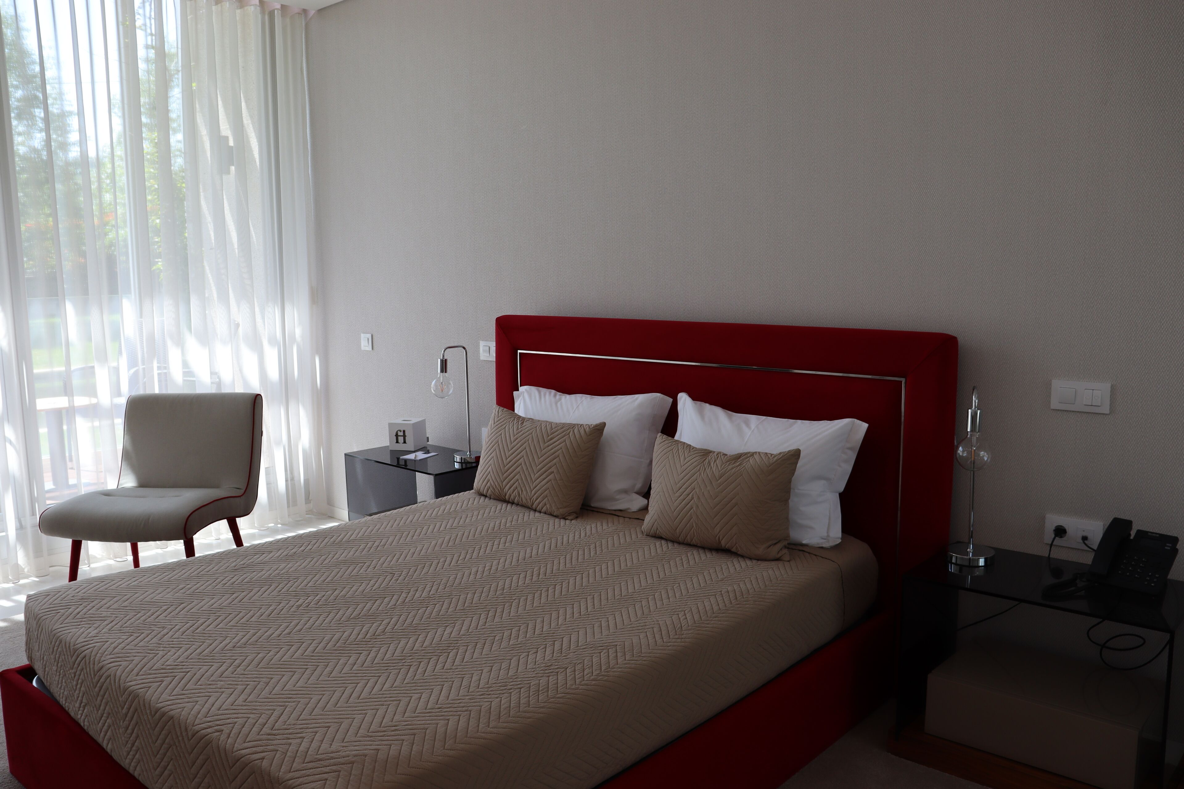 Una habitación de hotel con cabecera roja, una cama con ropa de cama color crema y blanca, una silla, una mesita de noche con lámpara y un teléfono.