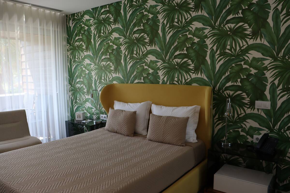 A hotel room with a bed, a chair, a nightstand, and a wall with a tropical leaf pattern.