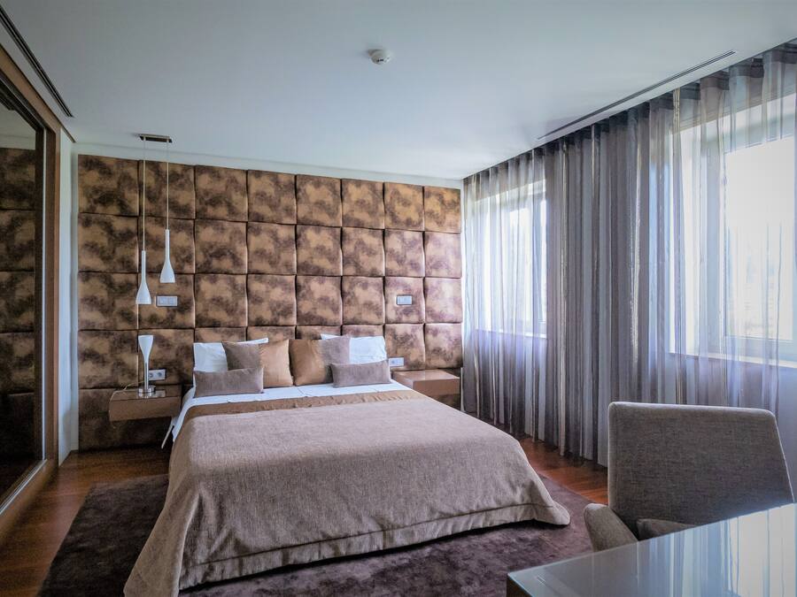 A modern hotel room with a large bed, a glass table, and a view of the city through sheer curtains.