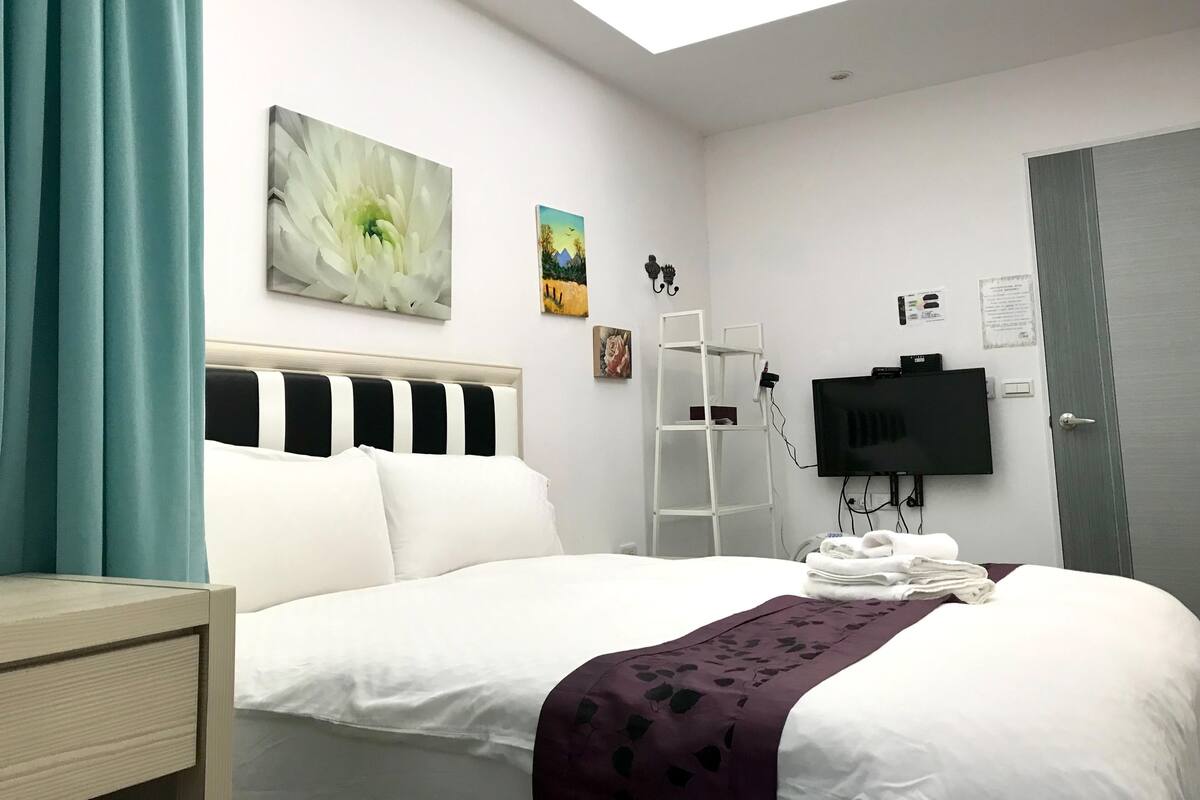 A hotel room with a bed, a TV, a painting, and a window.