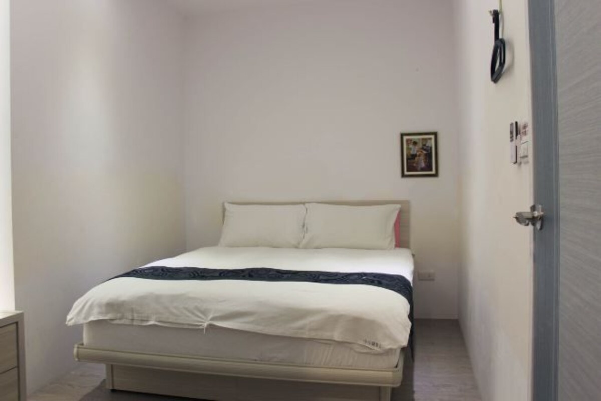 A narrow, single-bed room with a simple bed, a bedside table, and a door with a handle.