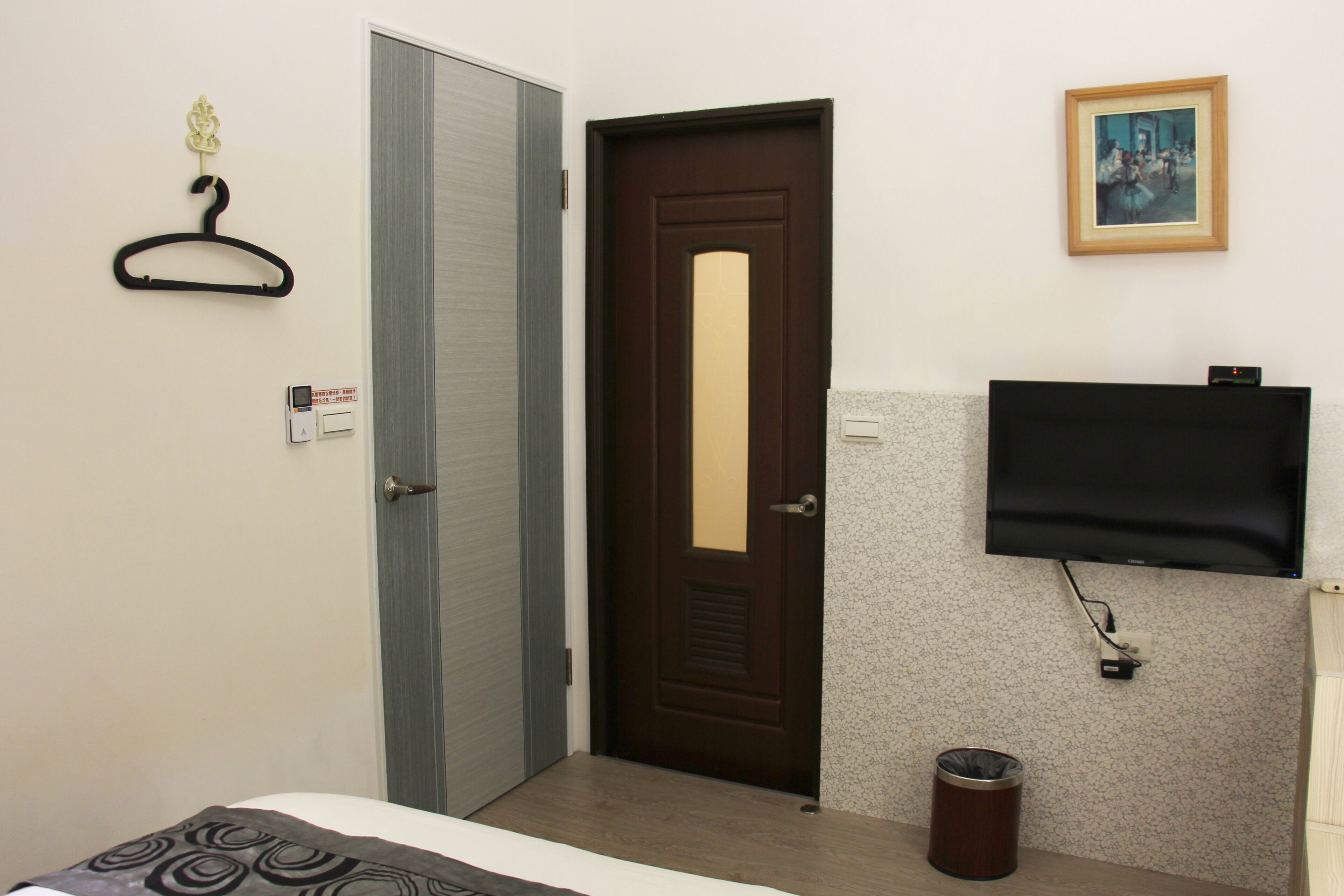 Hotellihuone, jossa on sänky, ovi, naulakko, roskakori ja seinälle asennettu televisio.