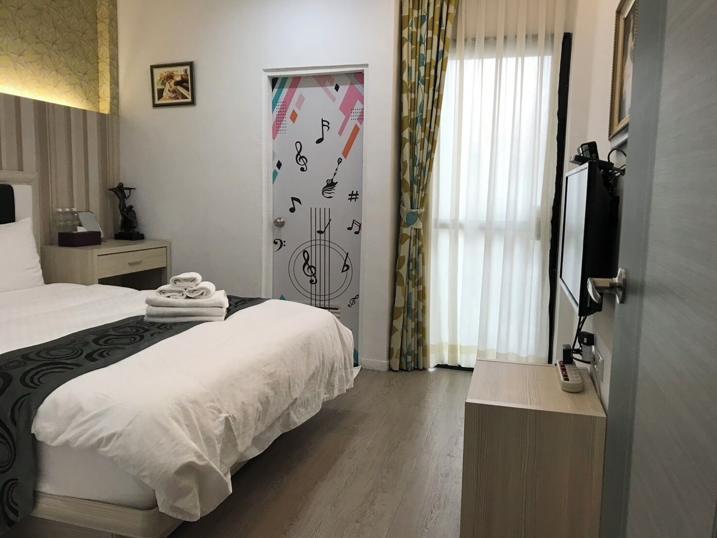 Une chambre d’hôtel avec un lit, une table de chevet avec une lampe, une télévision et un rideau orné d’un motif géométrique.