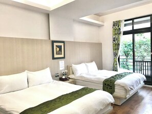 A hotel room with two beds, a framed picture on the wall, a window with a view of greenery, and a balcony.