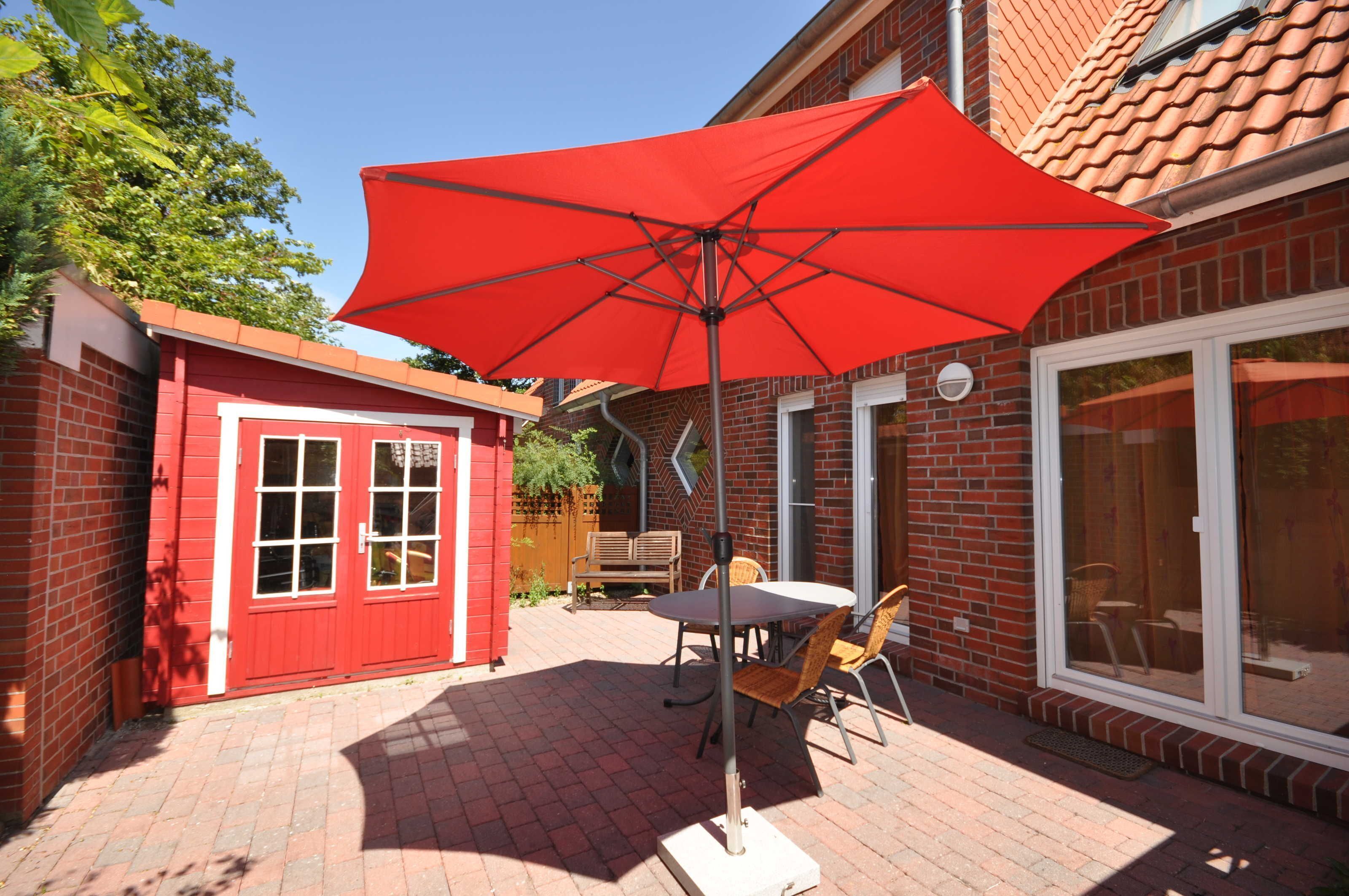 Un espace patio agrémenté d’un parasol rouge, d’un mur en briques, d’un petit abri de jardin rouge et d’une porte vitrée donnant sur un espace extérieur abrité.