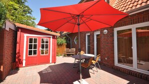 A patio area with a red umbrella, a brick wall, a small red shed, and a glass door leading to a covered outdoor space. - No. 39 - Cottage At the marina (Wittmund)